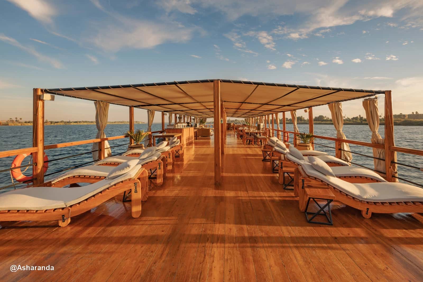 Deck Lounge – Dahabeya Nuun & Nut Cubierta lounge de la dahabeya Nuun & Nut con tumbonas de madera y toldo, navegando por el Nilo al atardecer.