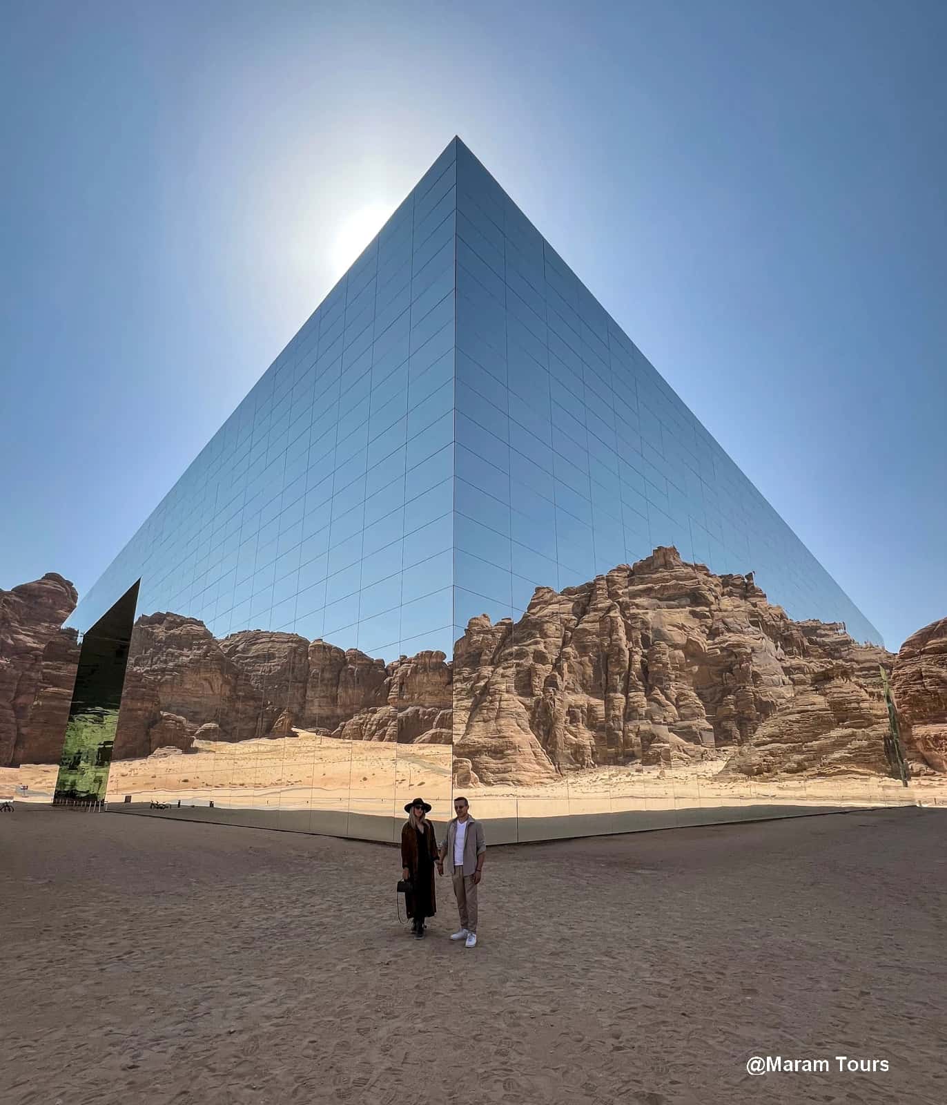 Maraya, el espejo del desierto en Al-‘Ula Edificio espejado en forma cúbica en el desierto de Al-‘Ula, Arabia Saudita, con una pareja de pie en primer plano y montañas rocosas reflejadas en su fachada.