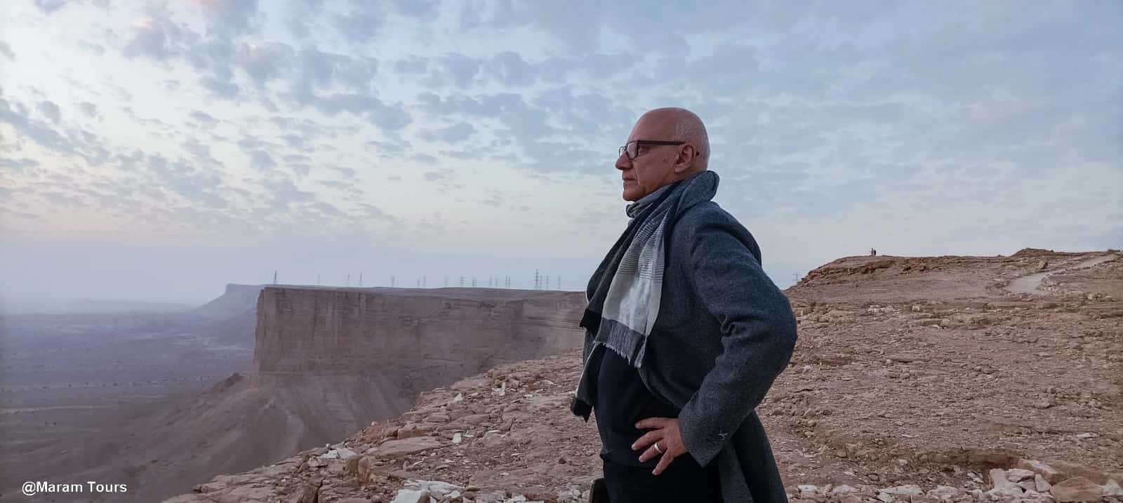 Explorando el Edge of the World – Arabia Saudita desde las alturas Hombre de pie sobre un acantilado rocoso llamado The Edge of the World en el desierto de Arabia Saudita al atardecer, mirando hacia el horizonte bajo un cielo parcialmente nublado.