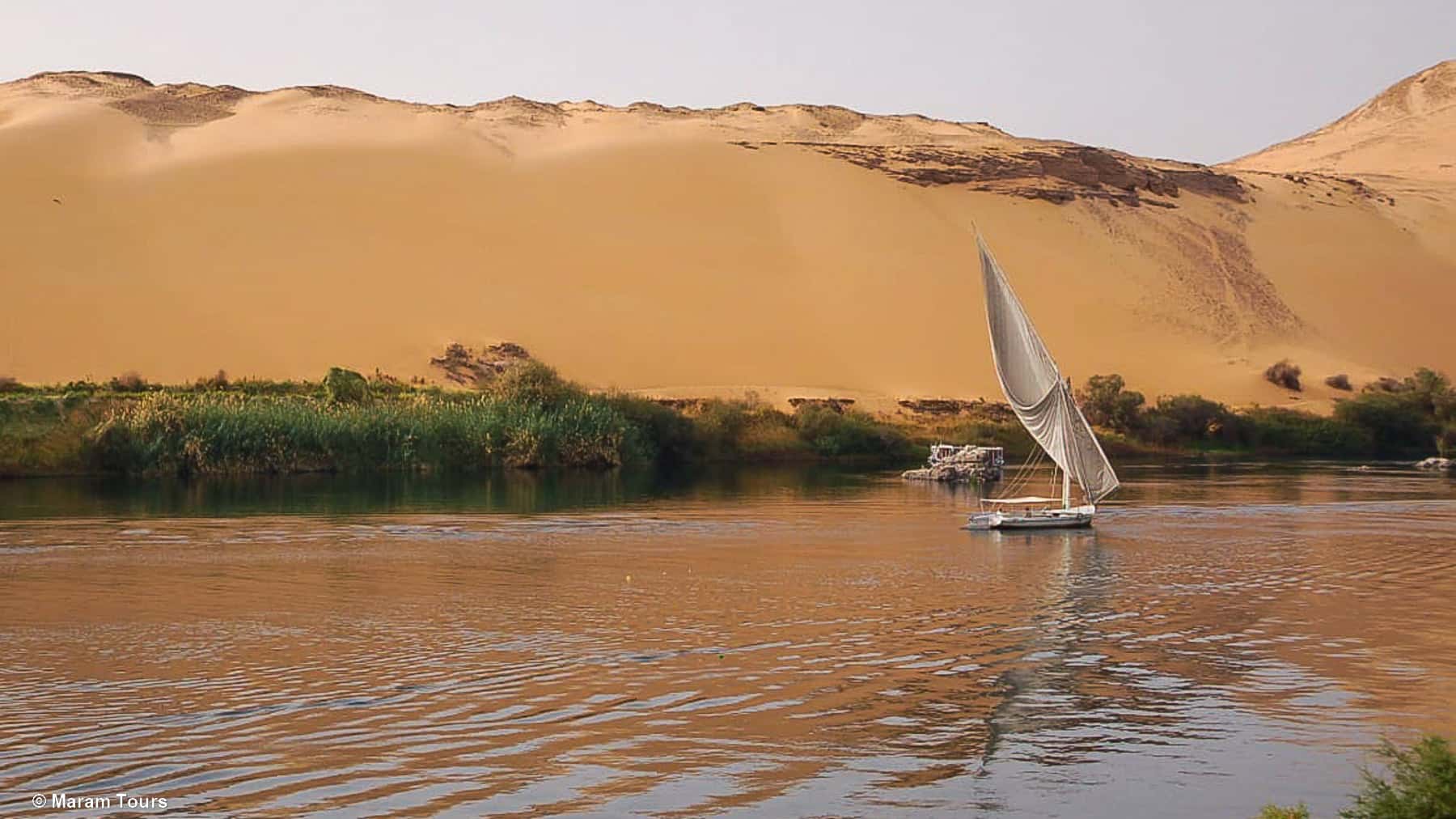 Egipto auténtico desde el agua Velero tradicional navega por el río Nilo al atardecer, frente a dunas doradas y vegetación densa en el sur de Egipto.